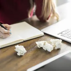 Osoba pisze w notatniku, tworząc wzór recenzji książki naukowej. Obok leży laptop i pogięte kartki.