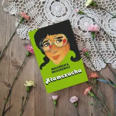 Recenzja książki "Kłamczucha" Małgorzaty Musierowicz. Okładka książki z rysunkiem dziewczynki w okularach, otoczona kwiatami na drewnianym tle.