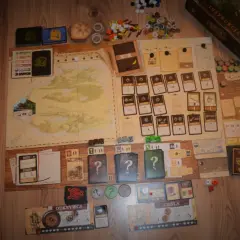Plansza gry planszowej "Robinson Crusoe" z kartami, żetonami i instrukcją. Recenzja książki Robinson Crusoe w tle.