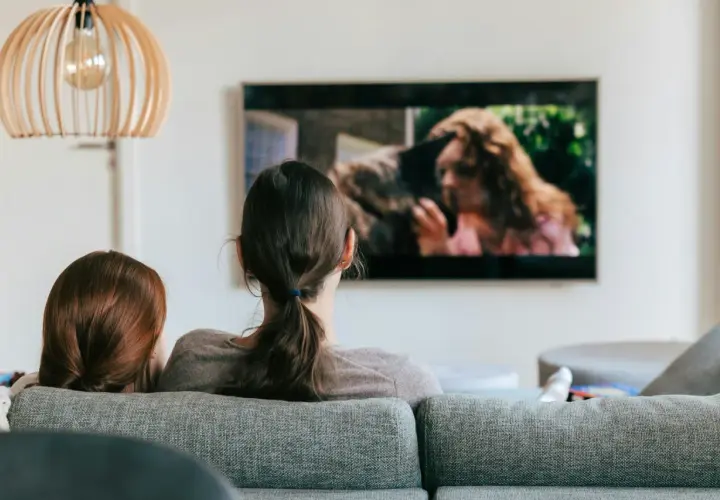 Dwie osoby oglądają film na ekranie telewizora. Na ekranie widać kobietę z rudymi włosami i zwierzę.