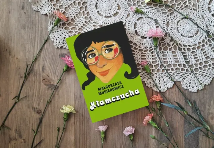 Recenzja książki "Kłamczucha" Małgorzaty Musierowicz. Okładka książki z rysunkiem dziewczynki w okularach, otoczona kwiatami na drewnianym tle.