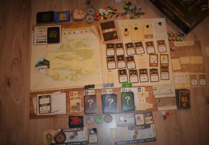 Plansza gry planszowej "Robinson Crusoe" z kartami, żetonami i instrukcją. Recenzja książki Robinson Crusoe w tle.