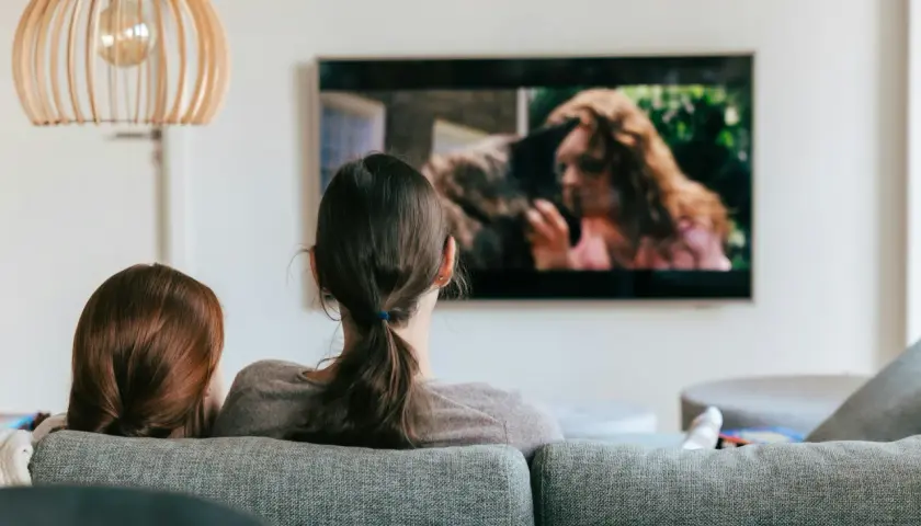 Dwie osoby oglądają film na ekranie telewizora. Na ekranie widać kobietę z rudymi włosami i zwierzę.