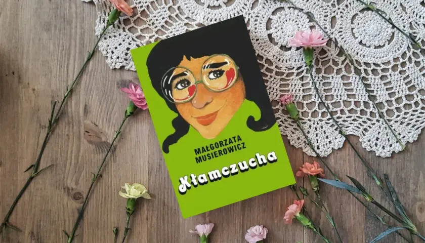 Recenzja książki "Kłamczucha" Małgorzaty Musierowicz. Okładka książki z rysunkiem dziewczynki w okularach, otoczona kwiatami na drewnianym tle.