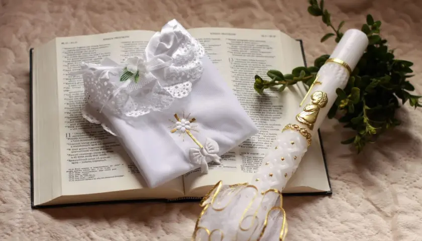 Biała szatka i paschalna świeca na otwartej Biblii. Symbole chrztu i ich znaczenie, światło wiary i czystość.