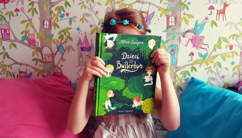 Dziecko z książką "Dzieci z Bullerbyn", której autorem jest Astrid Lindgren, na tle bajkowej tapety.