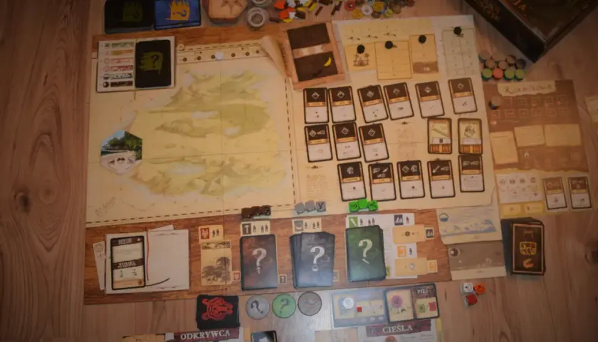 Plansza gry planszowej "Robinson Crusoe" z kartami, żetonami i instrukcją. Recenzja książki Robinson Crusoe w tle.