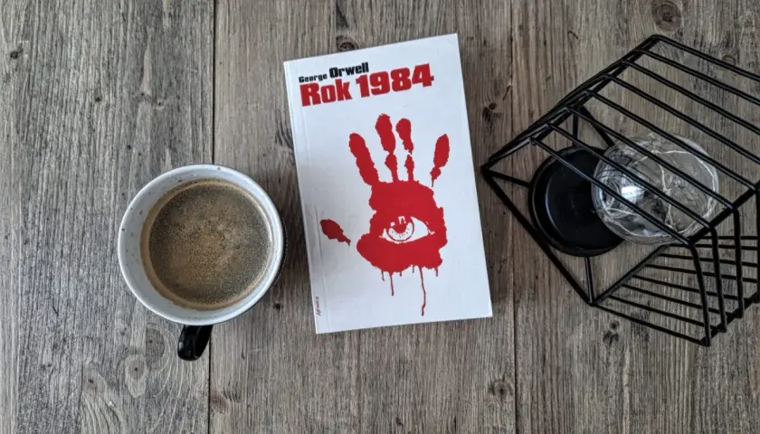Kawa i książka "Rok 1984" George'a Orwella na drewnianym stole. Recenzja tej ponadczasowej lektury czeka.
