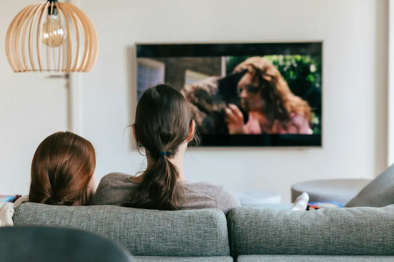 Dwie osoby oglądają film na ekranie telewizora. Na ekranie widać kobietę z rudymi włosami i zwierzę.