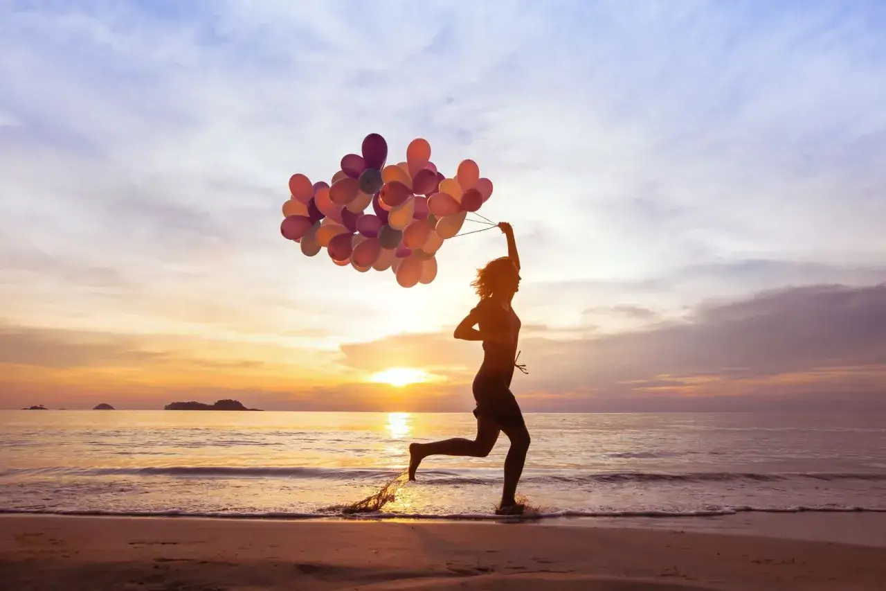 Sylwetka kobiety biegnącej z balonami na plaży o zachodzie słońca. Inspiracja do życia, jak w cytatach 